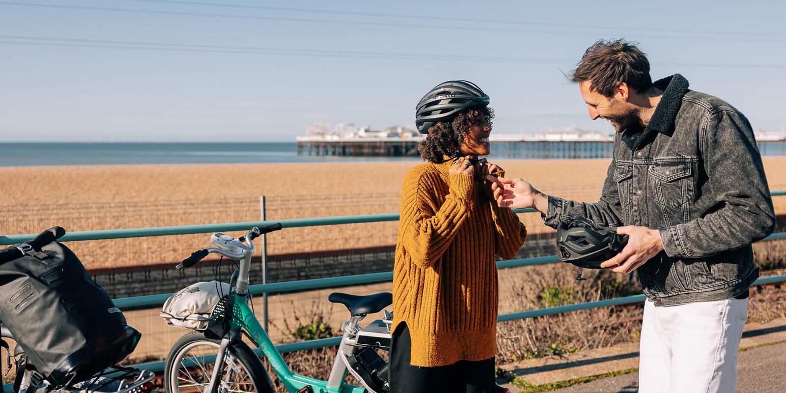 A man is helping a woman to adjust her bike helmet, they are standing in front of a beach with a pier
