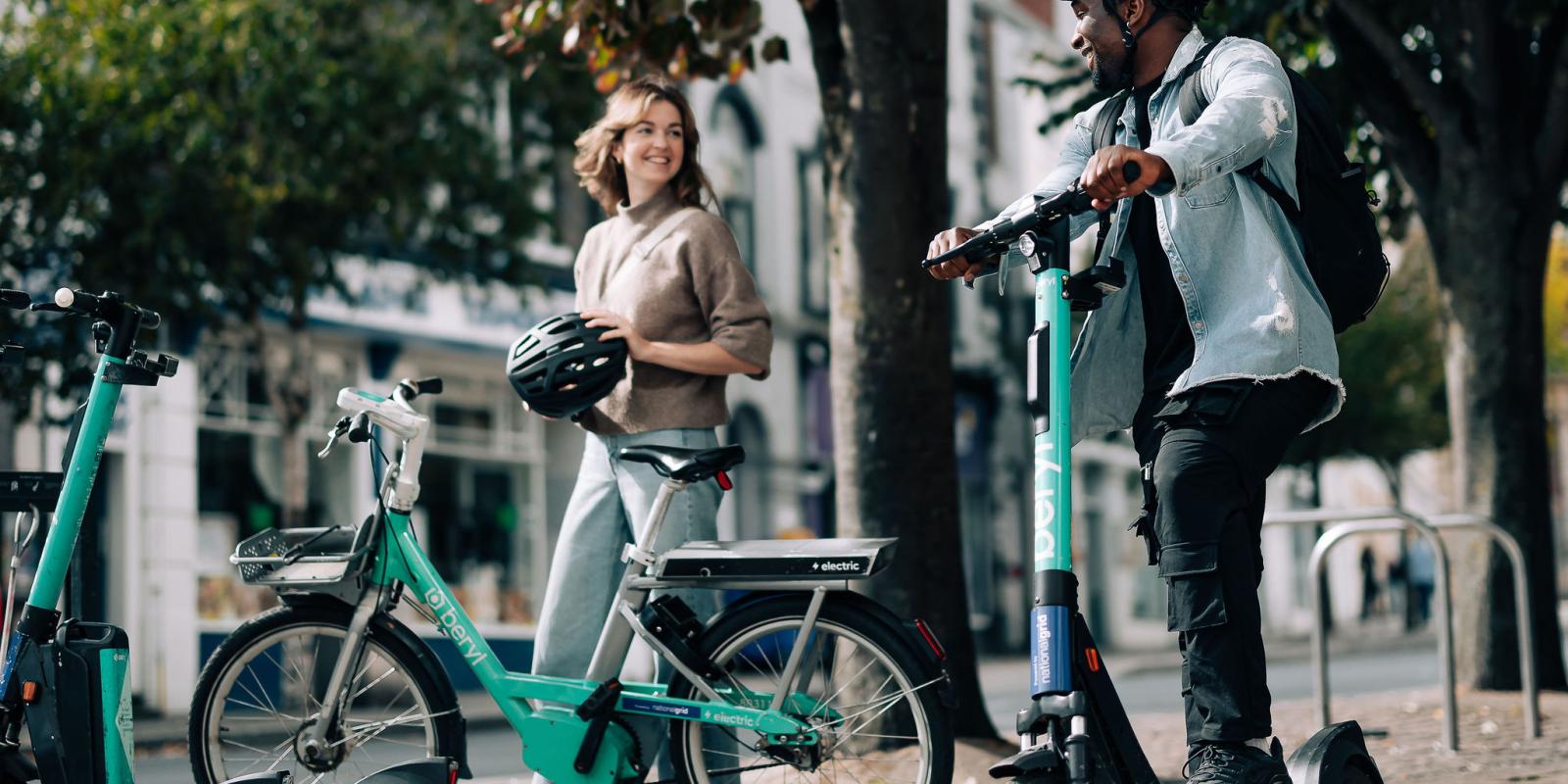 A woman is holding a helmet and stood next to a Beryl hire bike, she is smiling and looking at a man who has one foot on a Beryl e-scooter