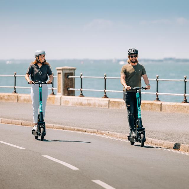 2 e-scooter riders on the road with the sea in the background 